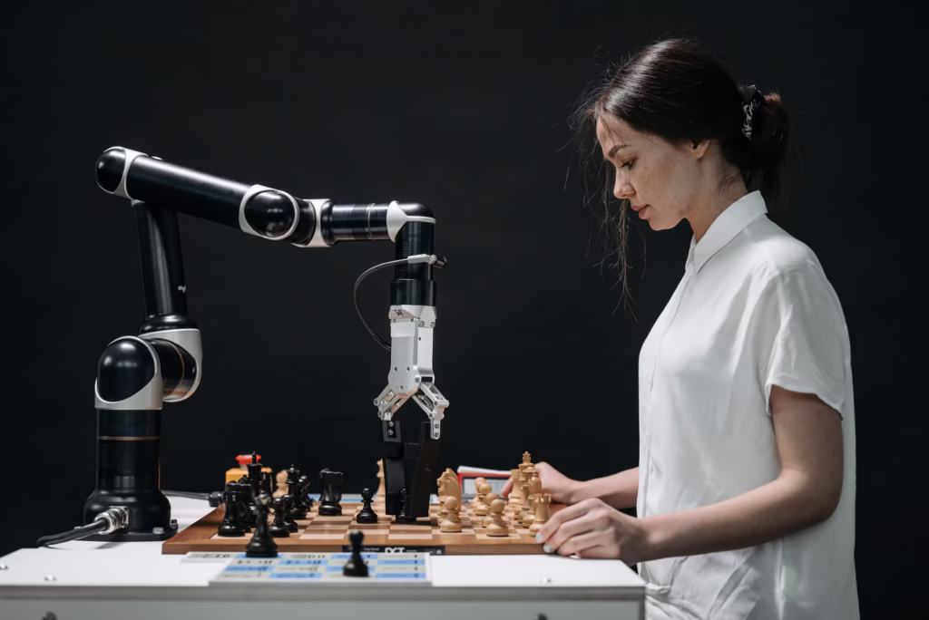 Białe ramię robota przemysłowego precyzyjnie przesuwające figurki na szachownicy obok siedzącej kobiety.